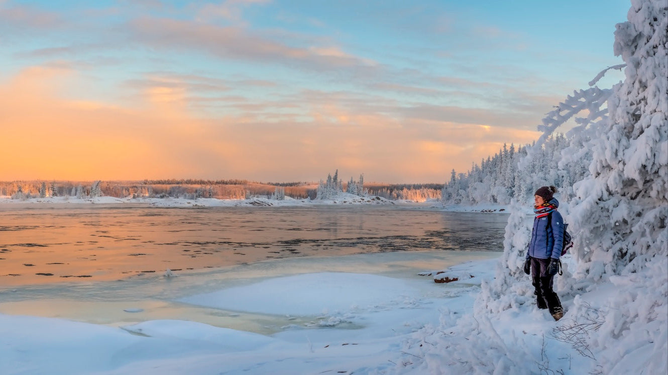 Person standing on a snowy landscape with a frozen river and sunset sky.