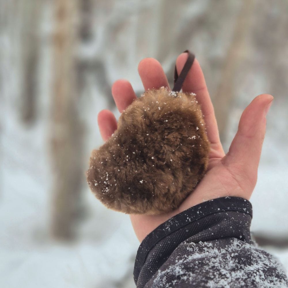 Hand holding a brown fur heart against a snowy forest background